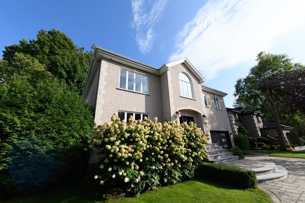 Stately two-story stucco home with arched entry, blooming hydrangeas, and circular paver driveway framed by mature trees.