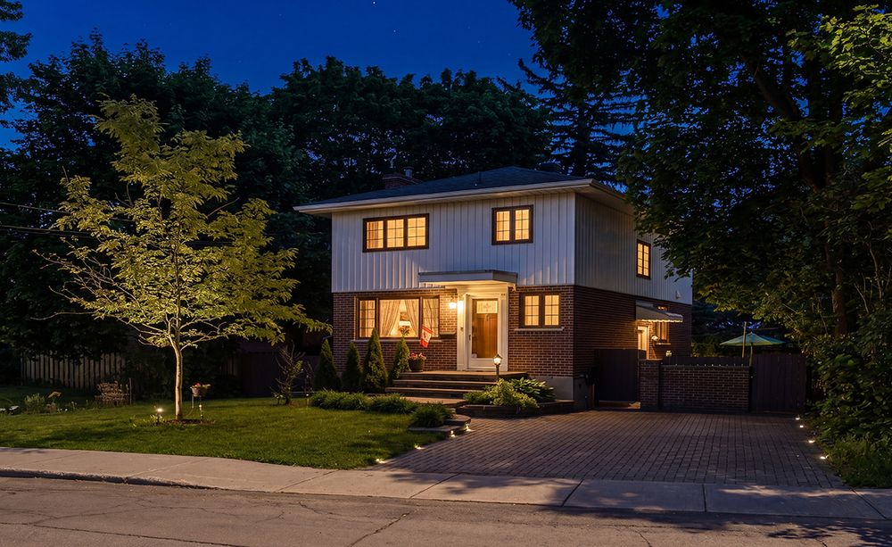 Modern two-story home with warm lighting at dusk, featuring white siding, manicured landscaping, and a paved driveway.