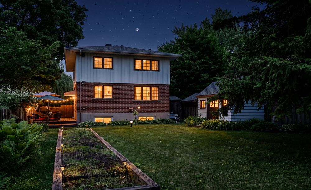 Two-story suburban home glowing warmly at twilight with lush green lawn and garden beds under a crescent moon.
