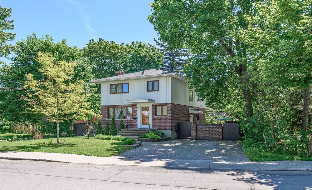 A two-story brick home surrounded by lush mature trees on a quiet suburban street in summer.