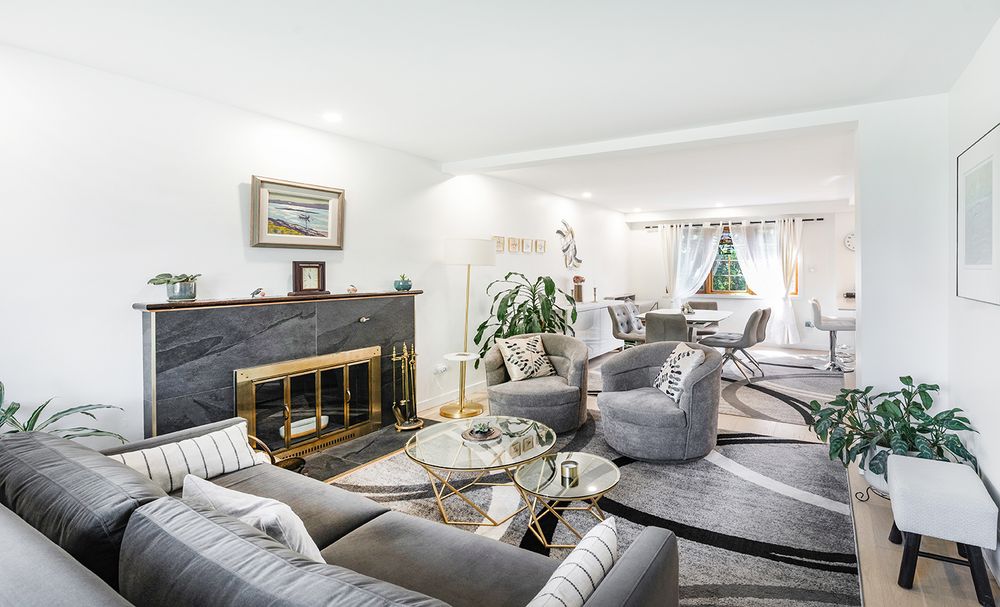 Bright living room with gray sectional sofa, marble fireplace, glass coffee table, and open dining area with natural light.