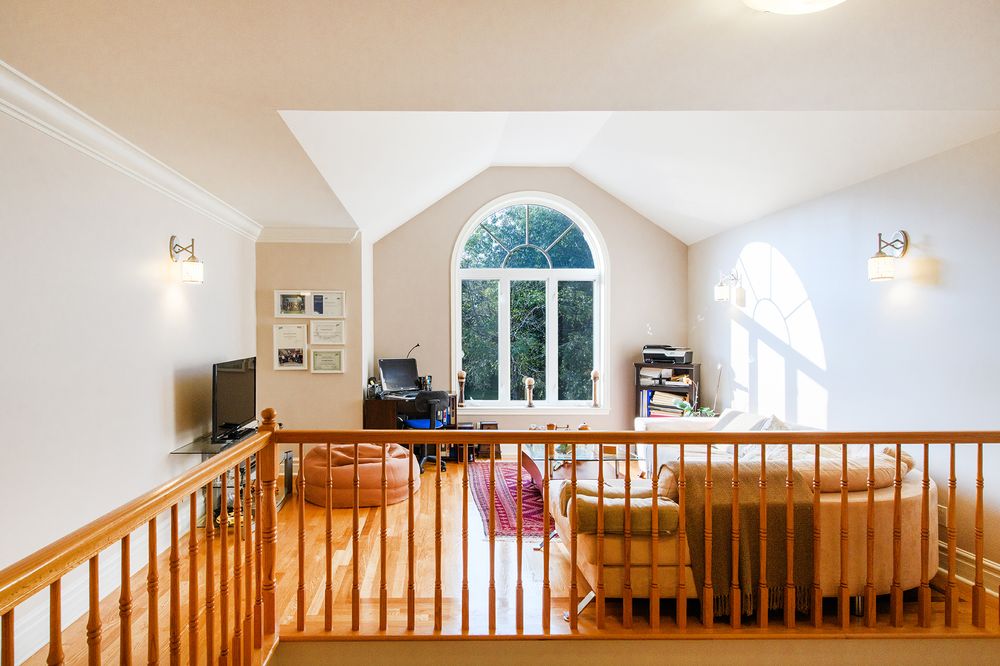 Bright upper-level loft area with hardwood railings, vaulted ceiling, arched window, and cozy seating area bathed in natural light.