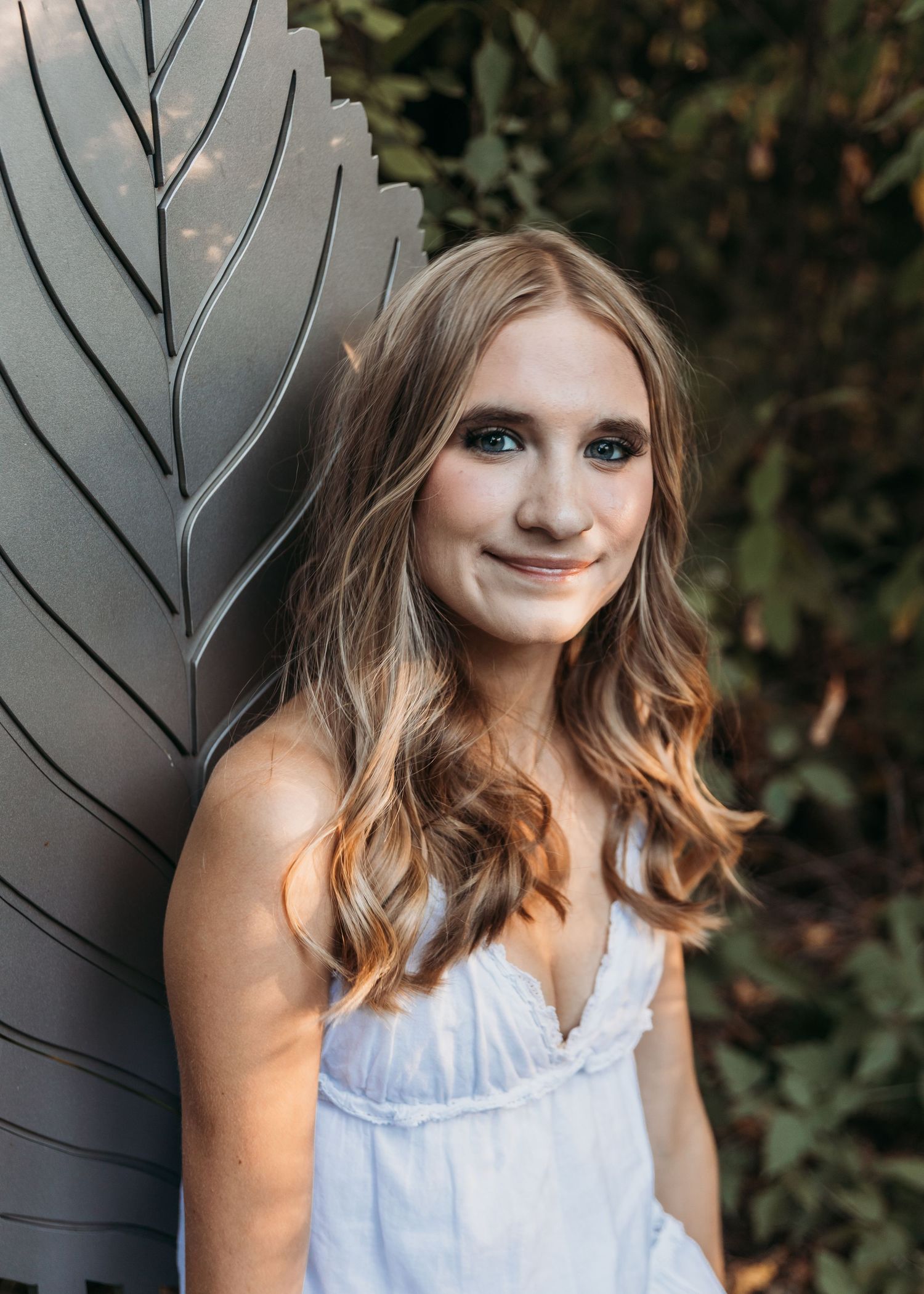 Young woman with long blonde hair smiles softly, wearing a white dress, posed against a decorative metal panel outdoors.