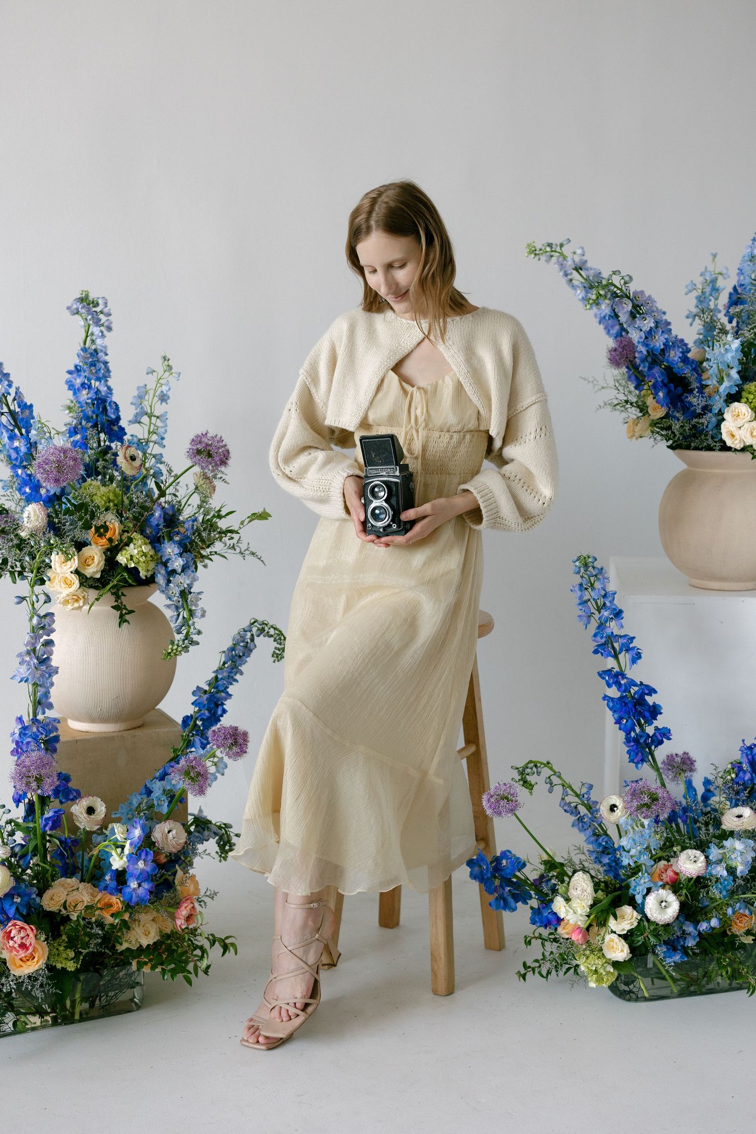 Woman in cream dress holding vintage camera surrounded by blue and white floral arrangements in a studio.