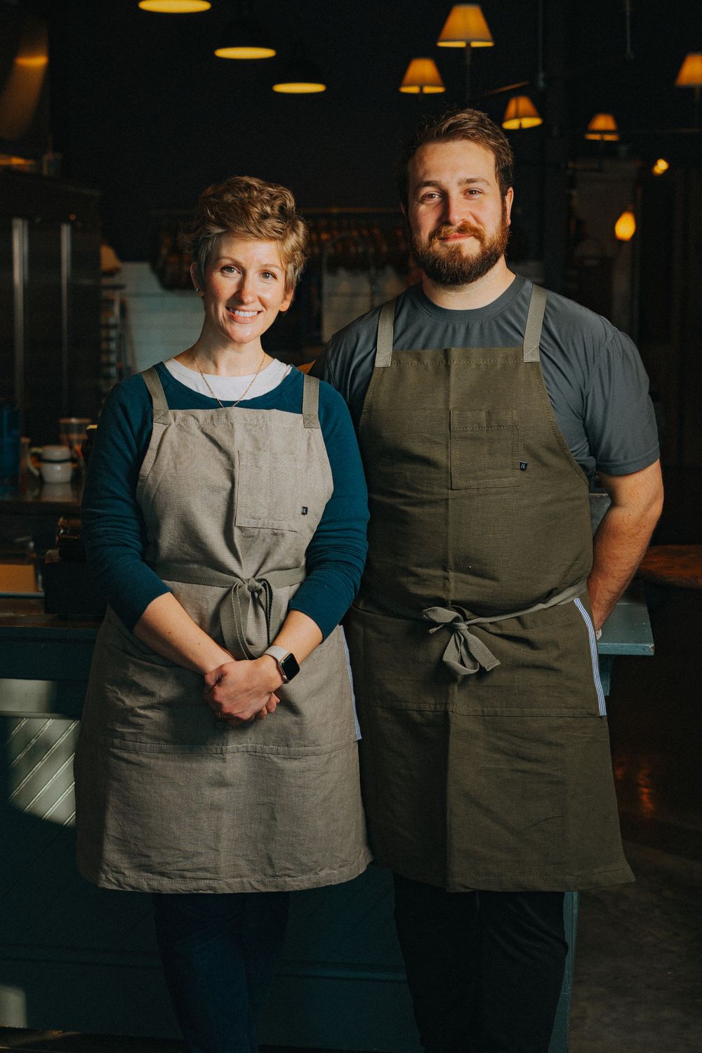 Two smiling restaurant workers wearing linen aprons pose together in a warmly lit professional kitchen.