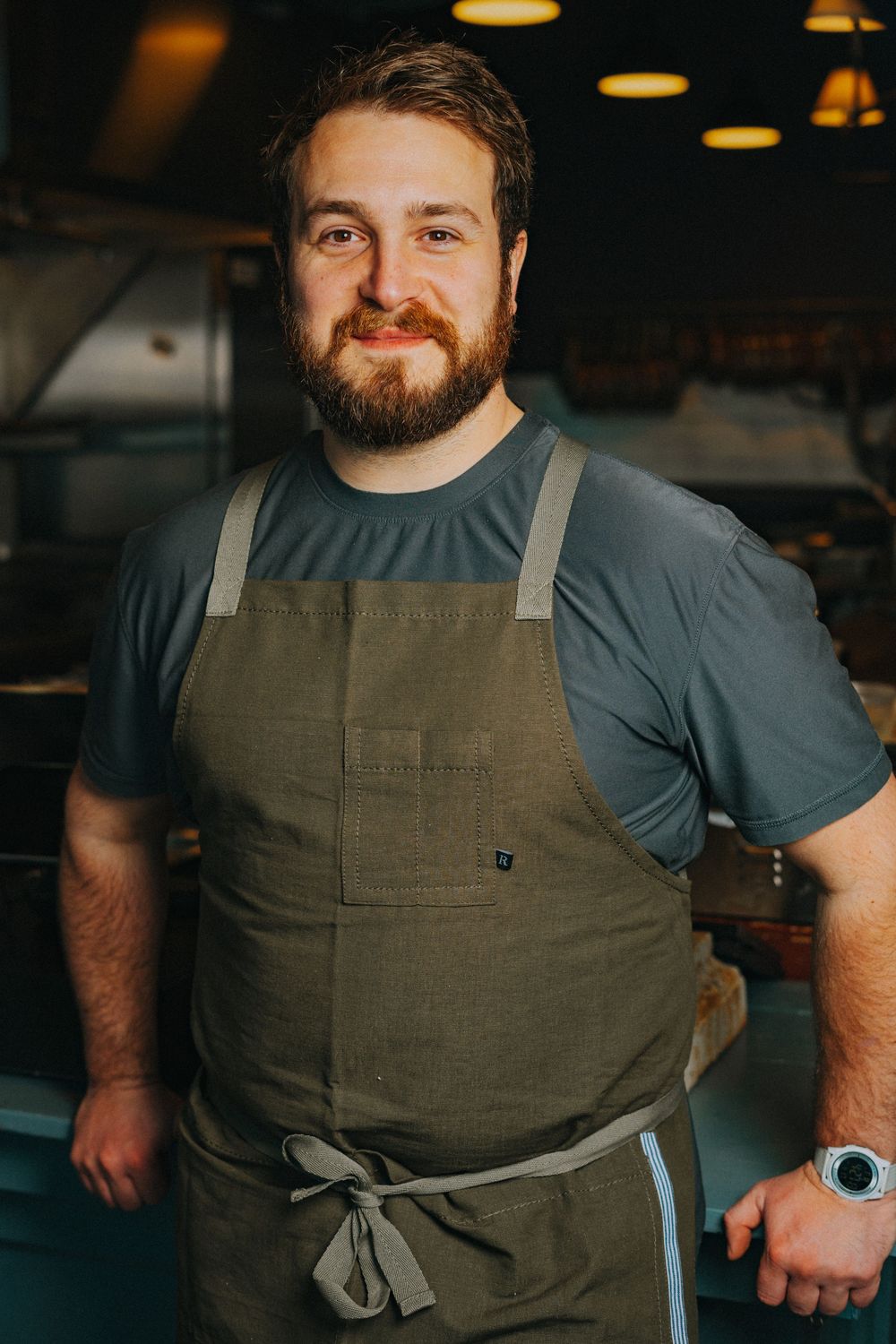 A bearded man wearing a gray shirt and olive apron smiles confidently in a professional restaurant kitchen.