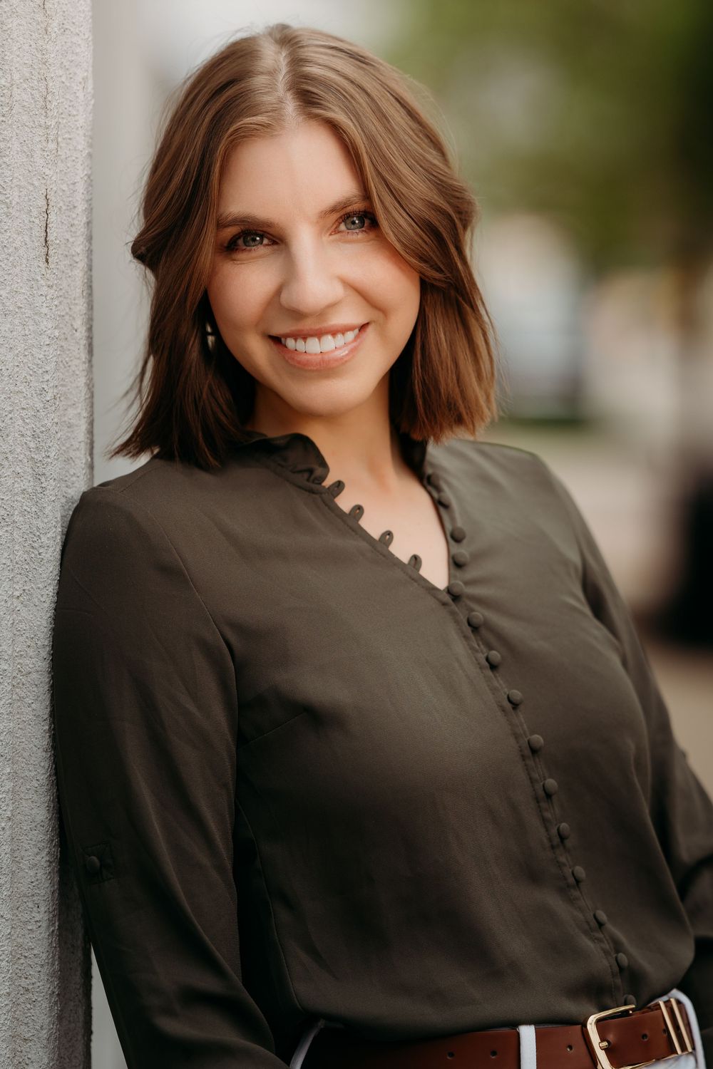 A smiling brunette woman with a bob haircut wears a dark olive blouse while leaning against an outdoor wall.