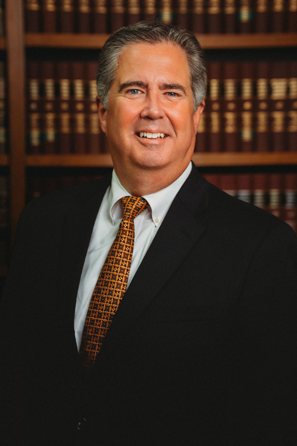 Professional male attorney in black suit with orange tie posing in front of law library bookshelves.