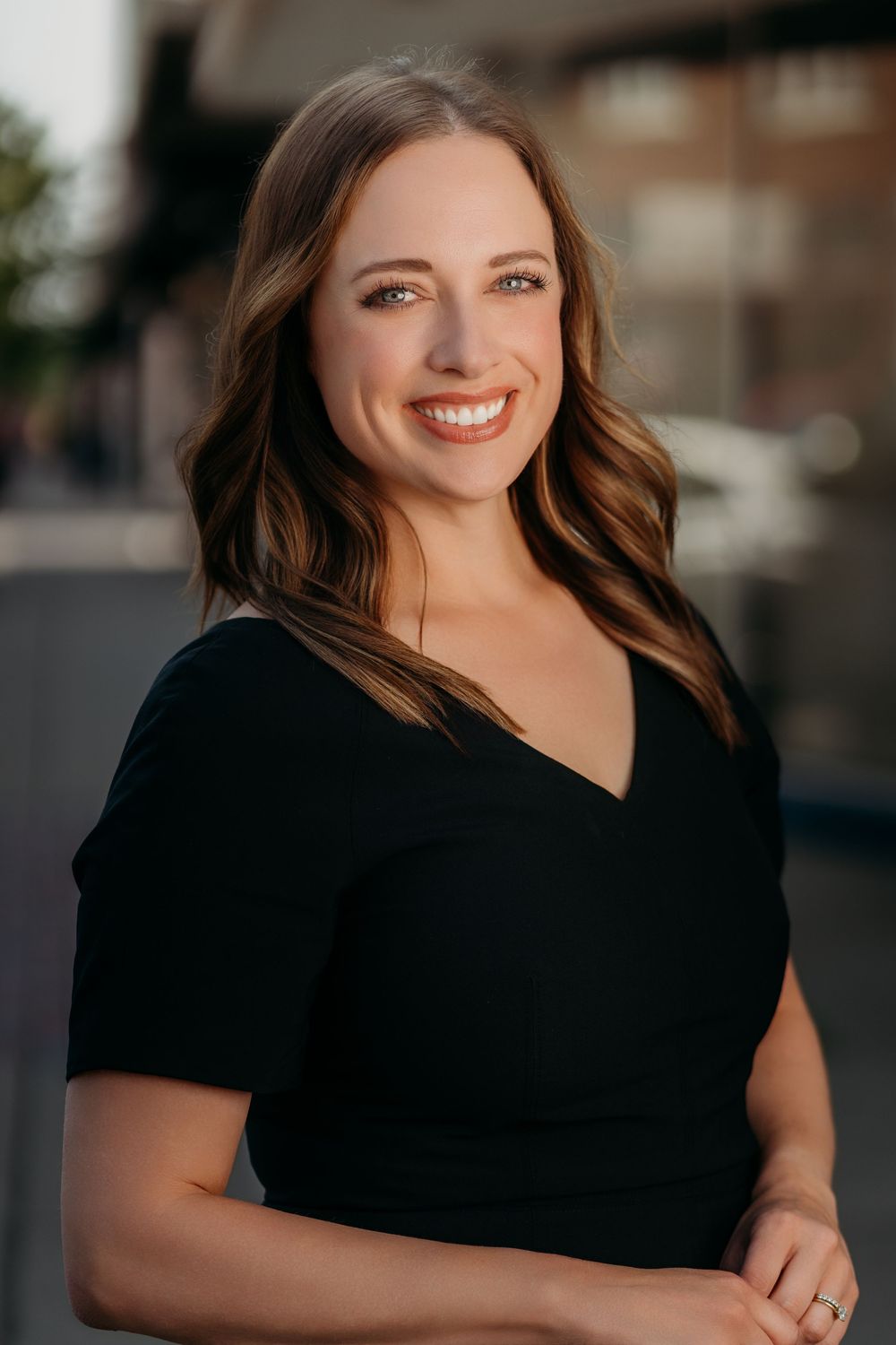 A smiling brunette woman in a black dress poses outdoors in a modern urban environment with soft bokeh background.