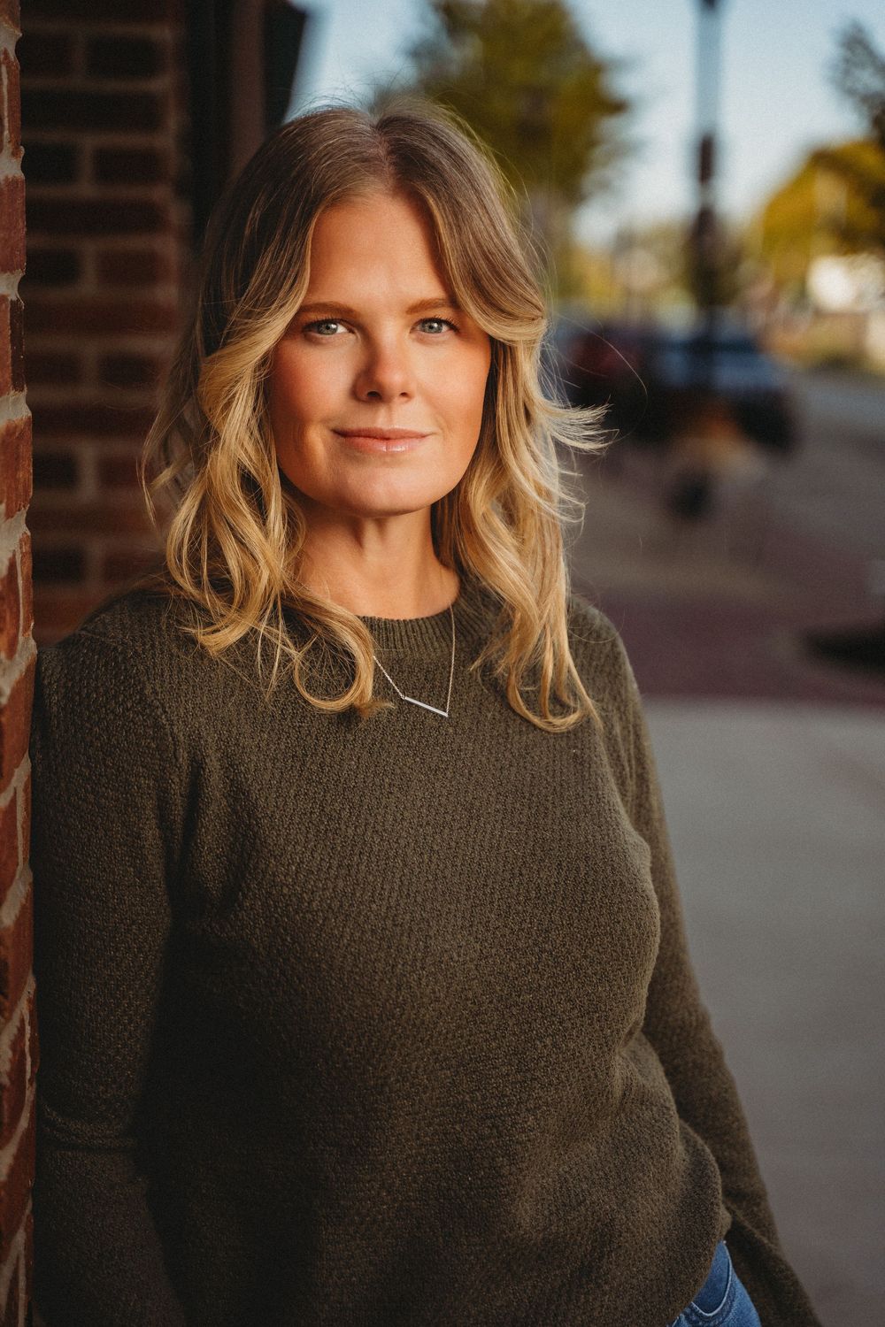 A blonde woman in an olive green sweater leans against a brick wall in warm golden hour light outdoors.