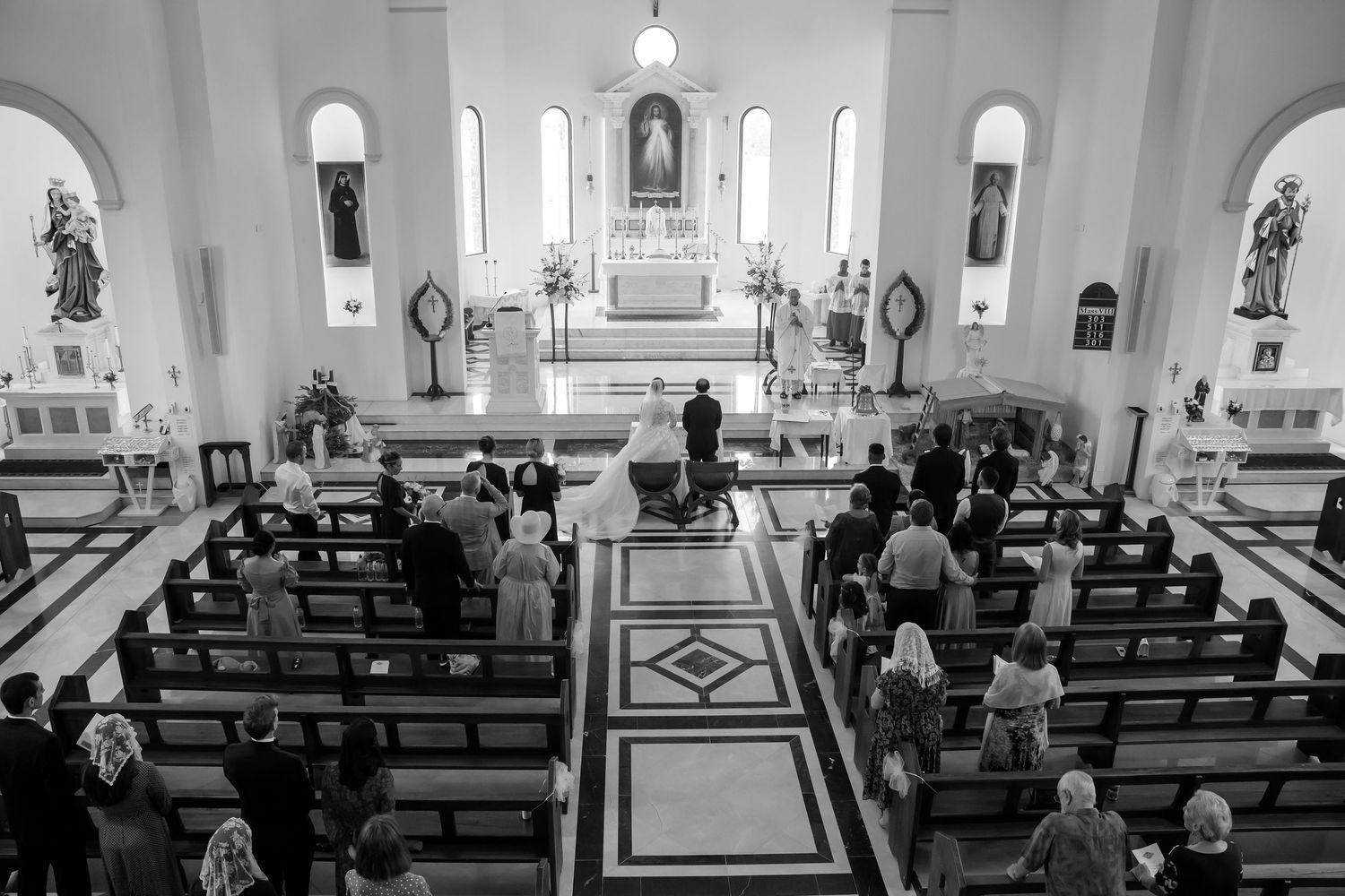 Black and white photo of wedding guests seated in church pews during a ceremony at a white altar.
