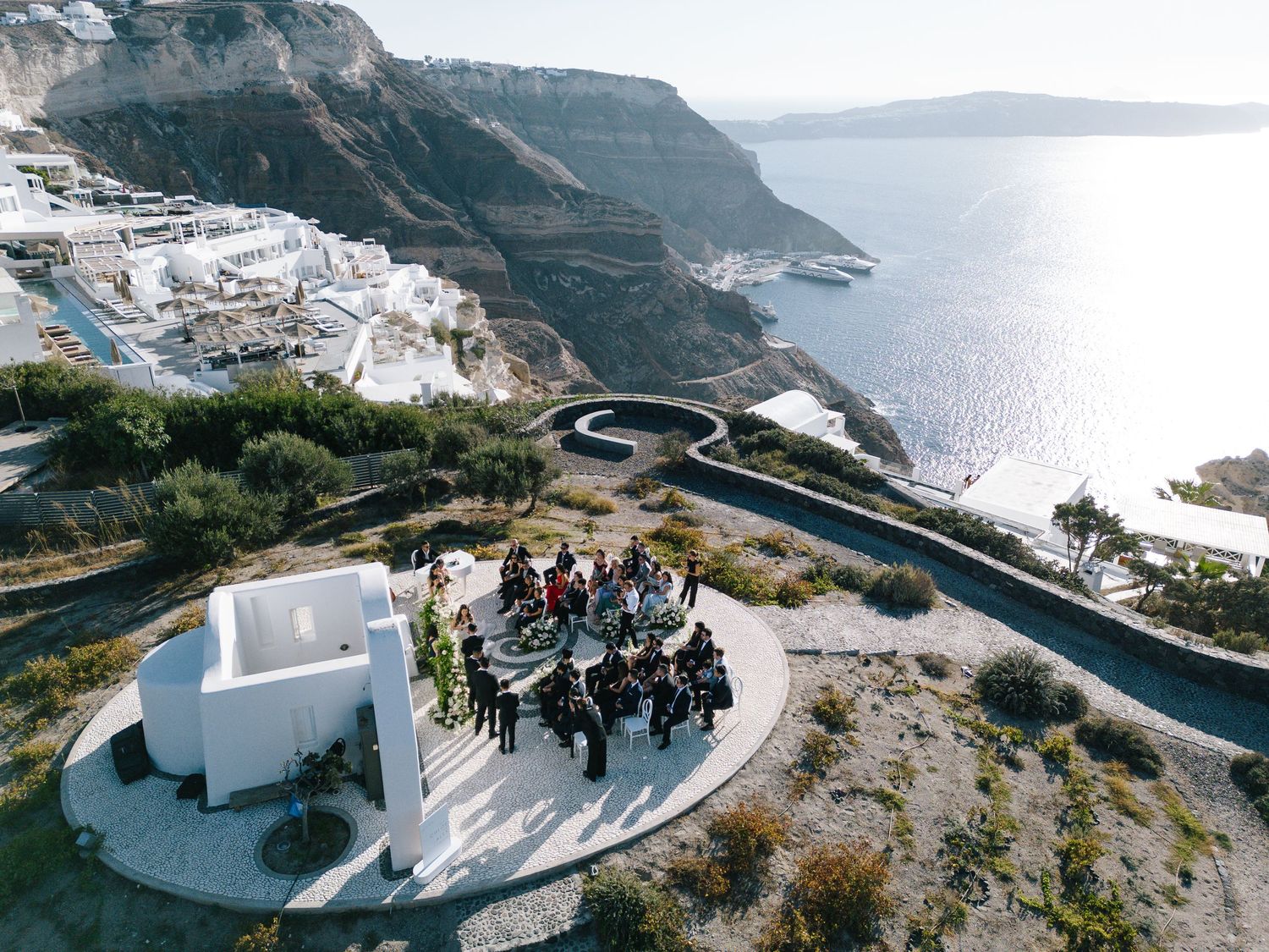 A person in a black suit stands at the edge of an infinity pool overlooking a scenic coastal view of Santorini.