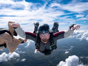 Two skydivers in freefall above clouds, wearing helmets and harnesses, with blue sky background.