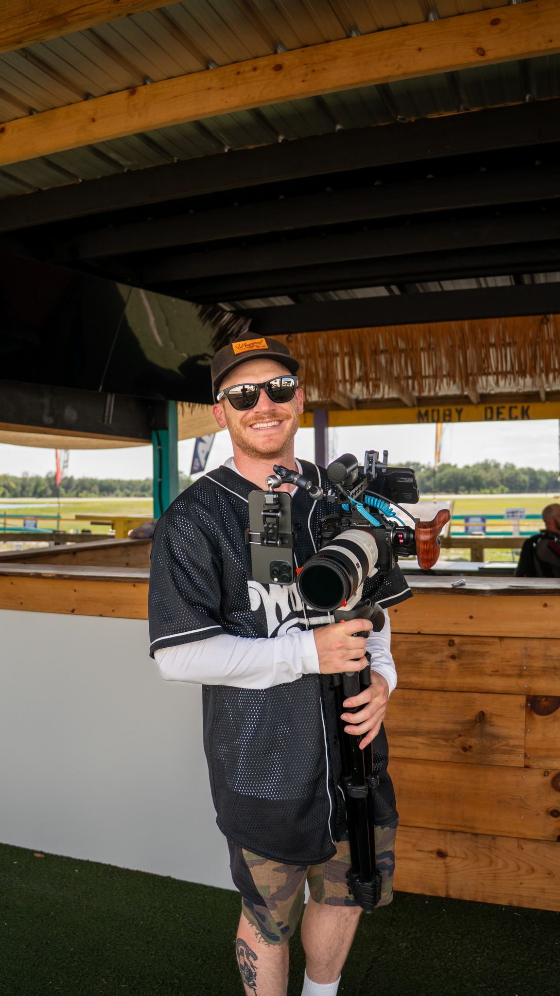 Smiling cameraman holding professional camera rig at outdoor Mojo Deck venue, wearing sunglasses and baseball jersey.