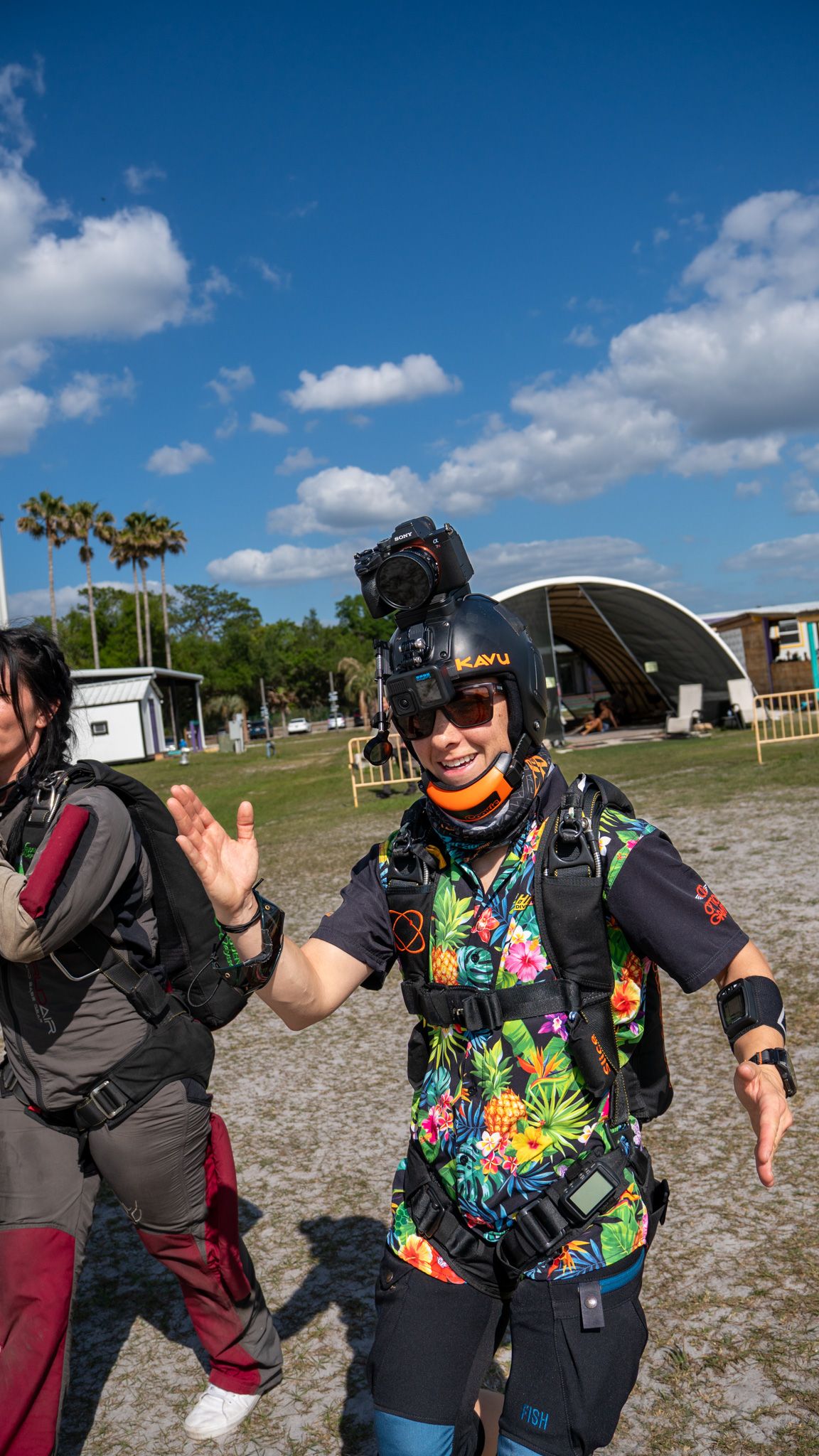 Smiling skydiver in colorful tropical jumpsuit with helmet camera waves after landing at a sunny dropzone.
