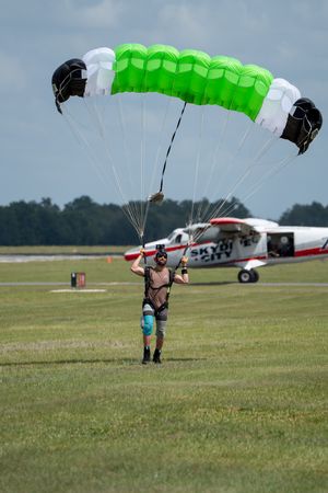 Skydiver landing with green parachute on airfield, Skydive City plane visible in background on sunny day.