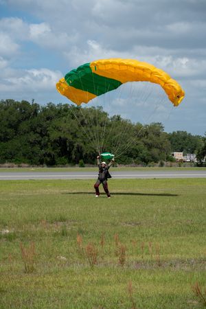 Skydiver landing on green grass field with yellow and green parachute canopy against cloudy blue sky.