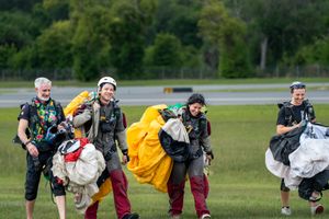 Four skydivers walk across a green airfield carrying colorful parachutes after a successful landing.