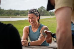 Smiling tattooed woman in blue tank top laughing at outdoor picnic table.