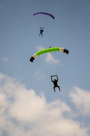 Two skydivers descend with colorful parachutes, one green and one purple, against a partly cloudy blue sky.