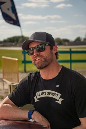 Man in black Leaps of Hope t-shirt and cap smiling outdoors at a racetrack on a sunny day.