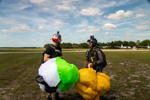 Two skydivers pack colorful parachutes on a grassy airfield after landing.