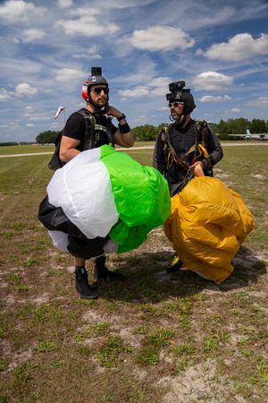 Two skydivers on a grassy airfield hold bundled green, white, and yellow parachutes after a successful landing.