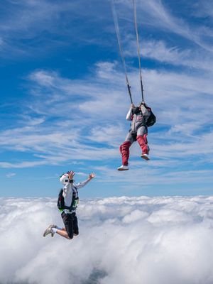Two skydivers perform aerial maneuvers above a thick cloud layer, one suspended by parachute lines against a vivid blue sky.