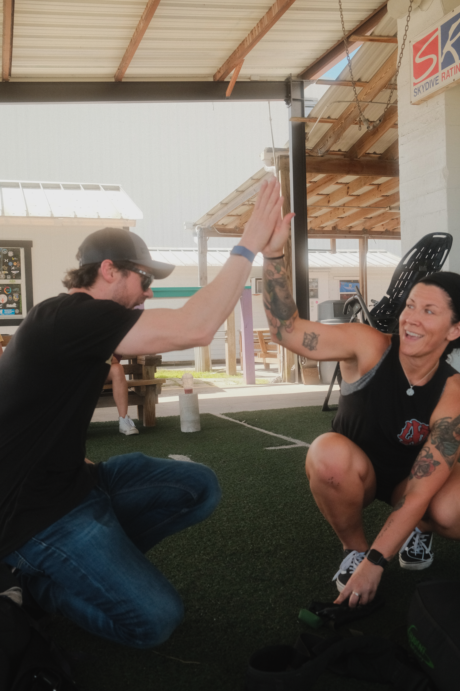 Two people high-fiving while kneeling on turf at an outdoor fitness event, both wearing black athletic wear.