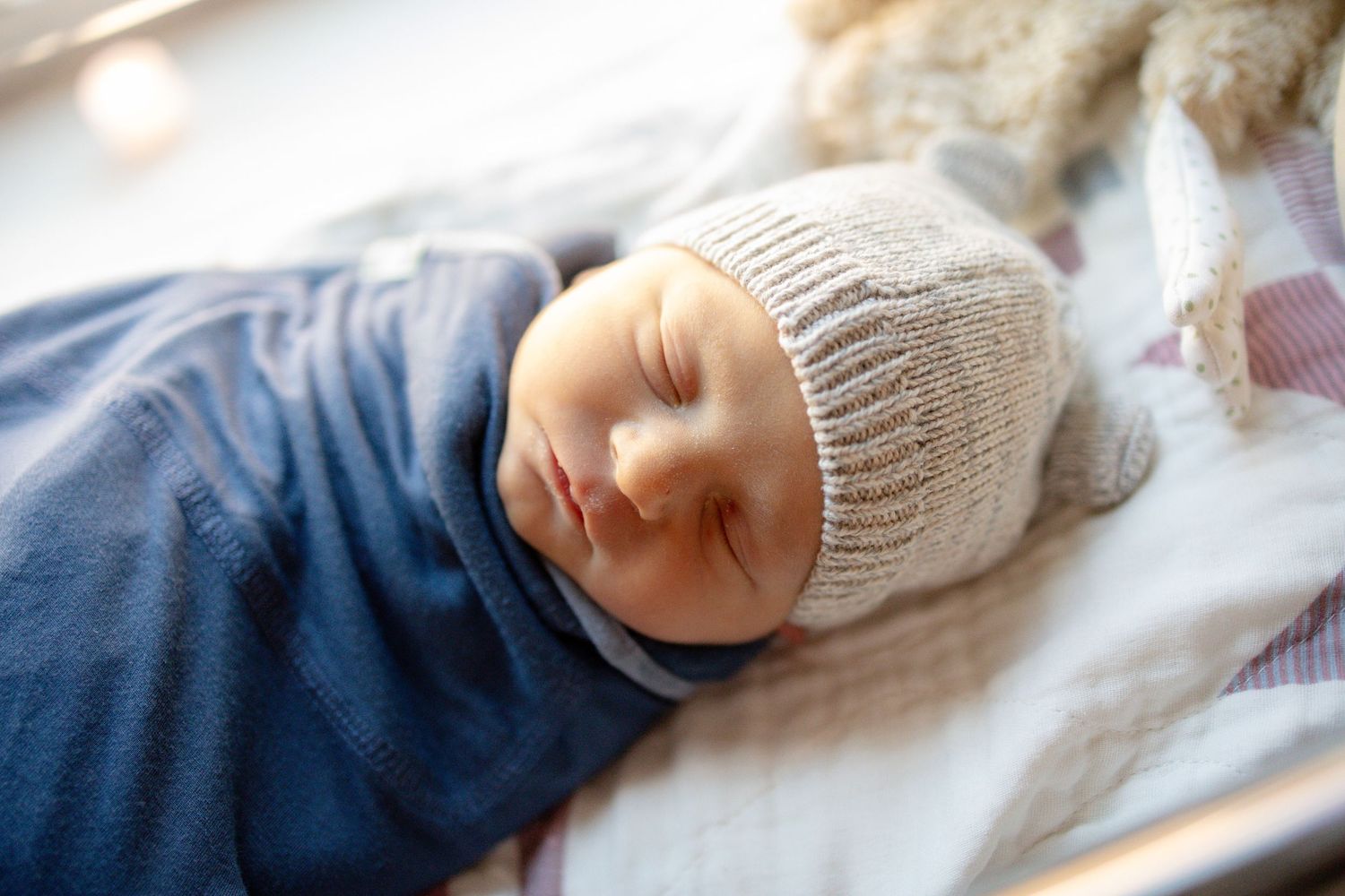 Peaceful newborn baby sleeping swaddled in blue wrap wearing a knit beige hat in a hospital bassinet.
