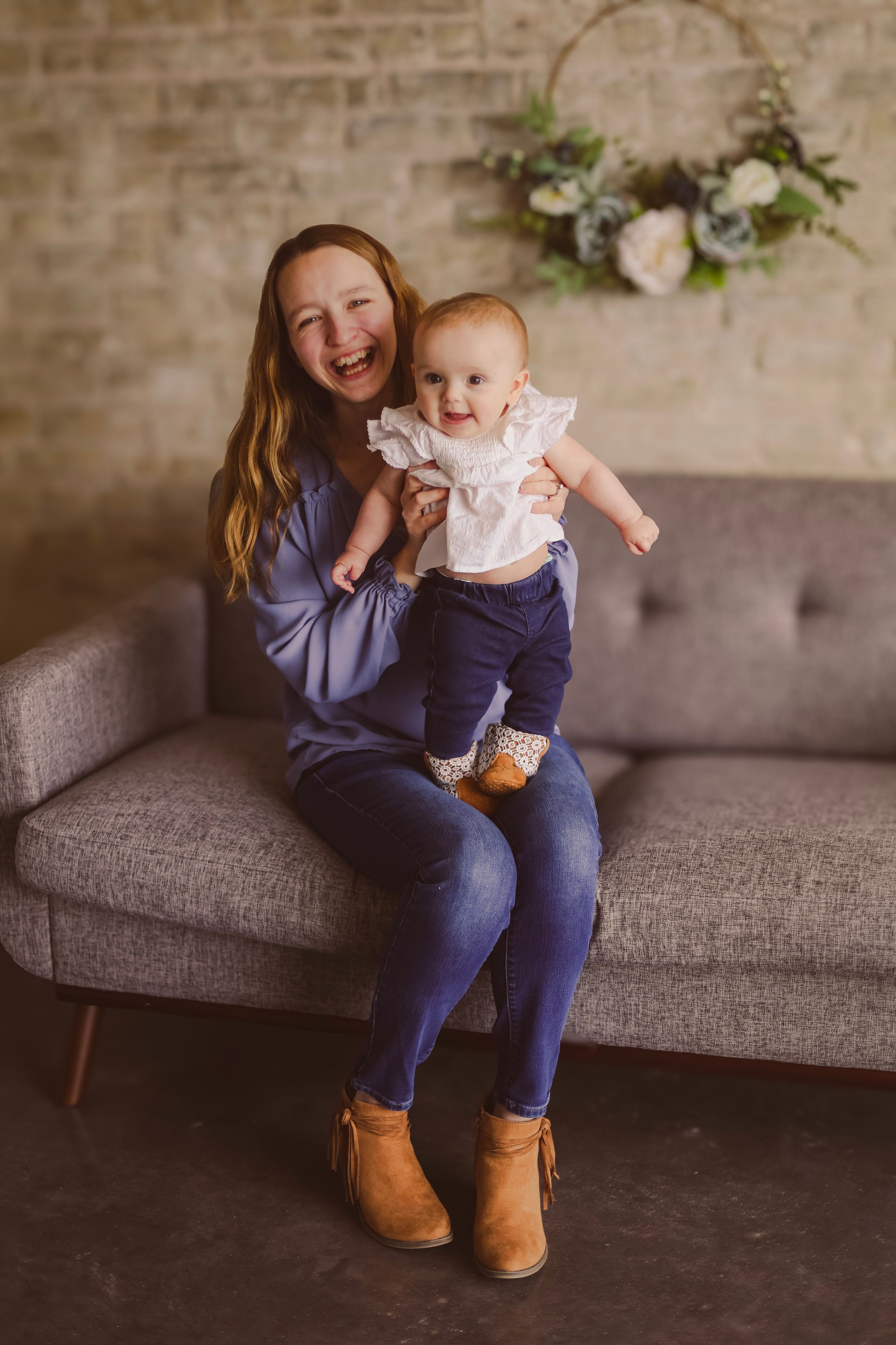 Laughing girl holding a baby on a gray sofa with a floral hoop wreath on a brick wall backdrop.