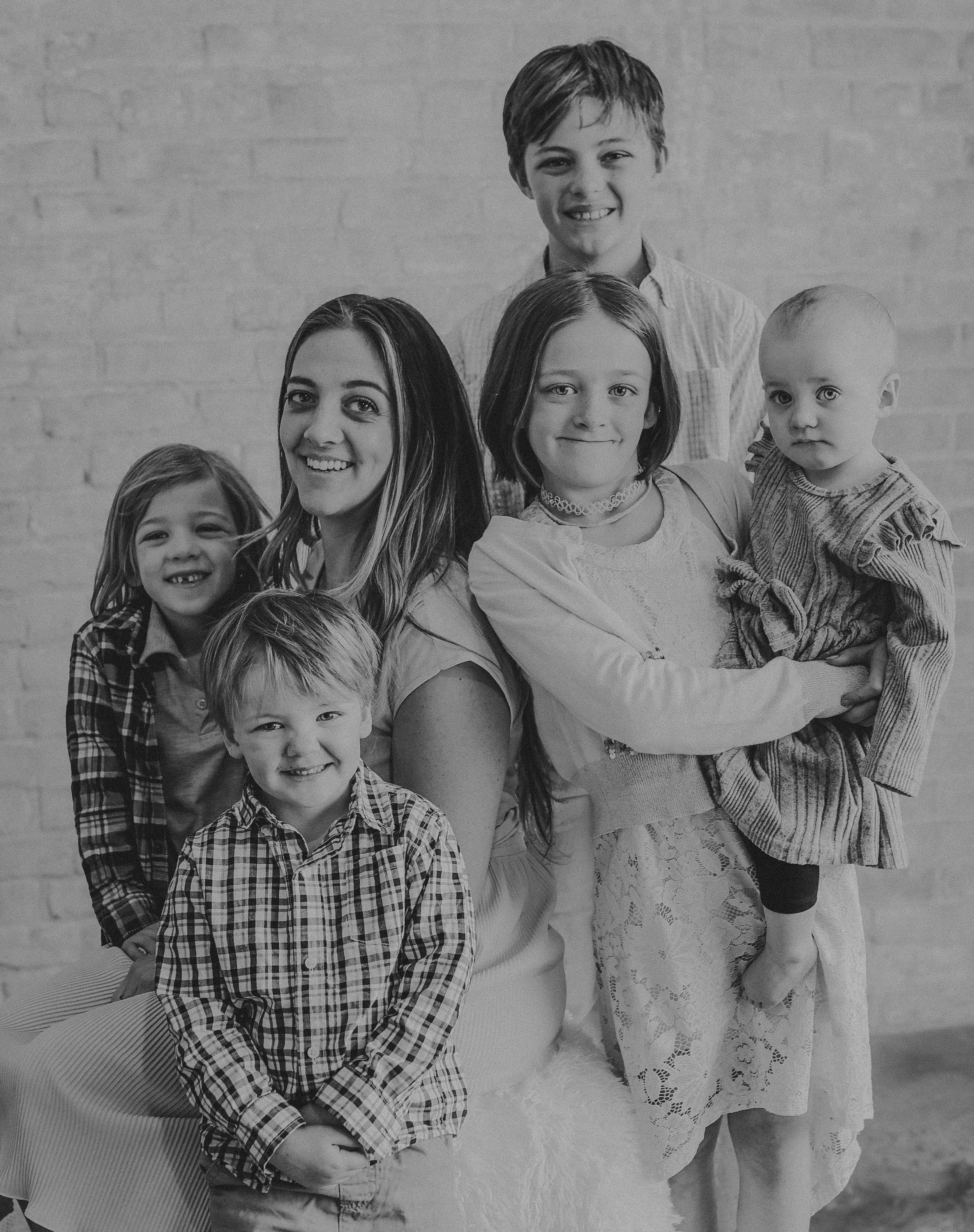 Black and white family portrait of a young woman with five children smiling together against a brick wall.