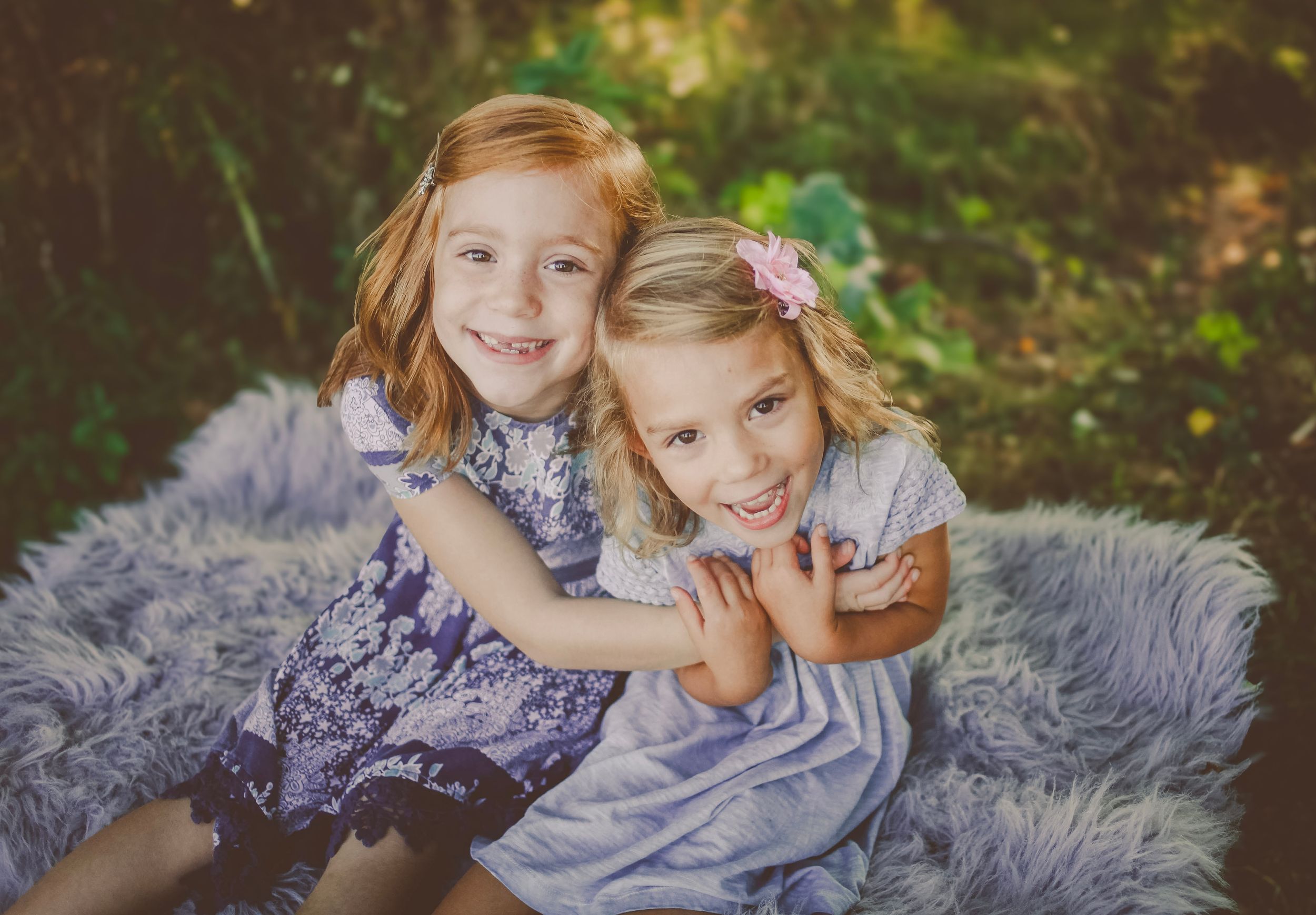 Two smiling young girls in blue dresses sitting on a fluffy gray rug outdoors in a garden setting.