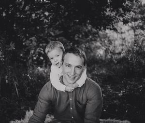 Black and white photo of a smiling man with a young toddler on his shoulders outdoors.