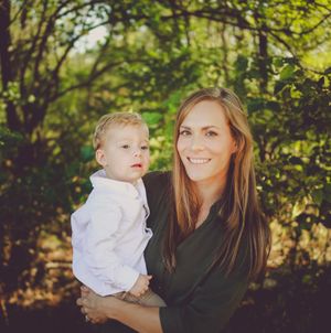 A smiling woman with long brown hair holds a young blonde toddler boy outdoors among lush green trees.