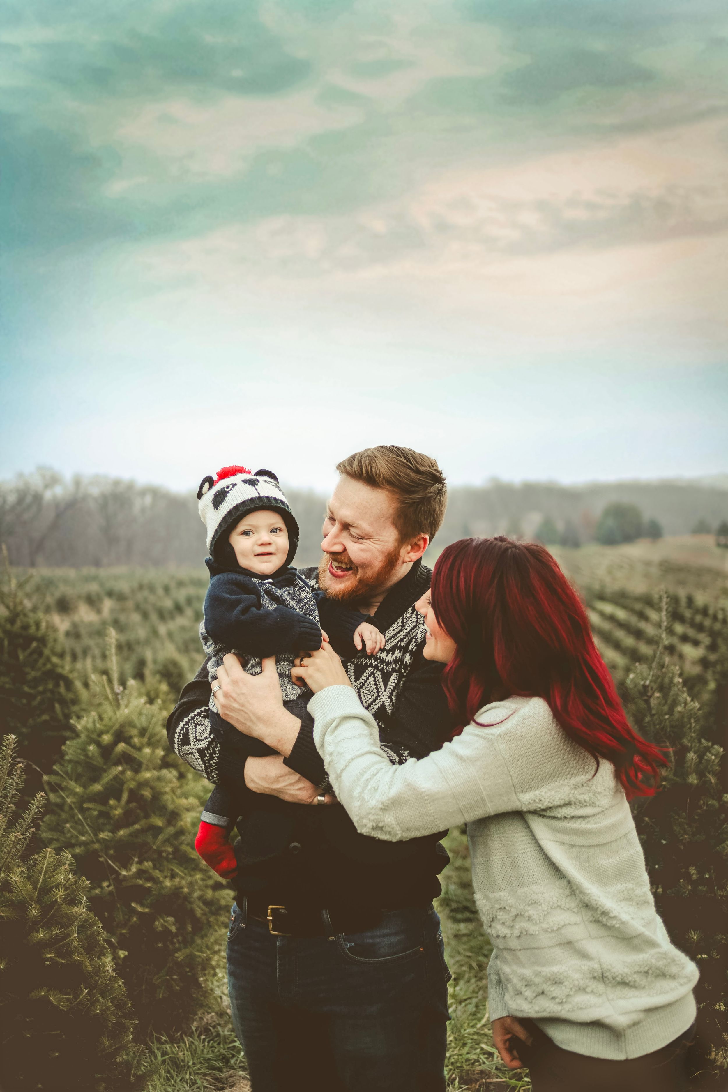 A young family with a bearded father holding a baby in a holiday hat, mother with red hair, at a Christmas tree farm.