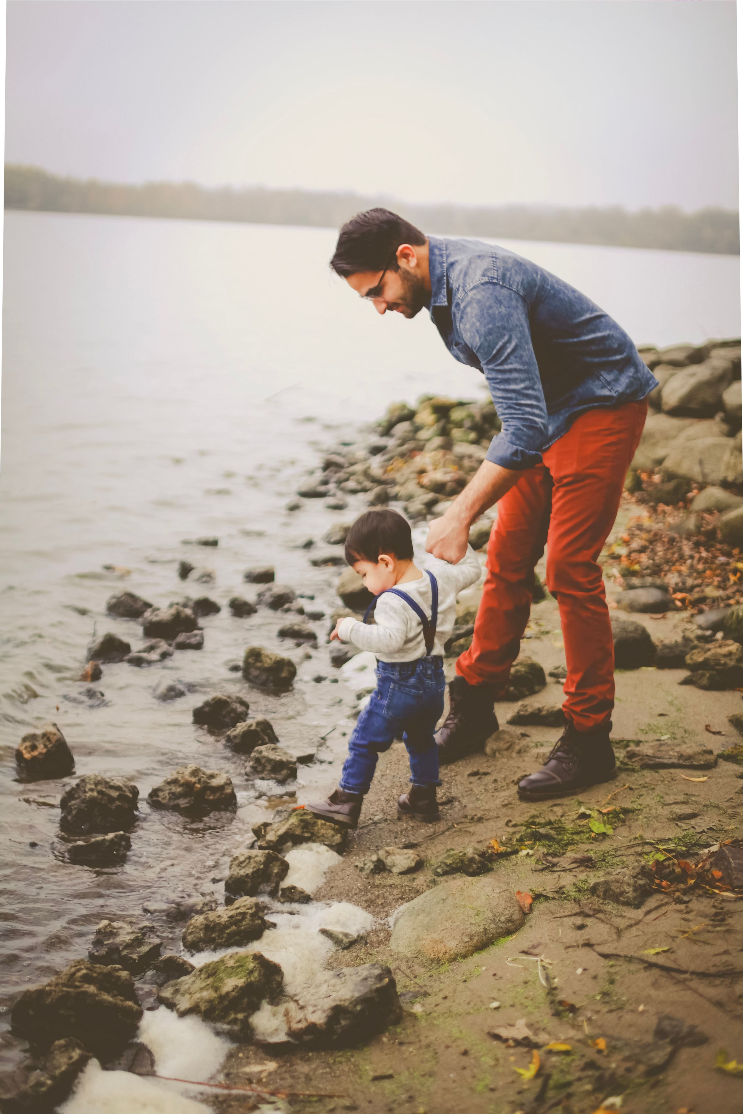 Father helping toddler walk on rocky shoreline near calm lake, both dressed in casual autumn clothing.