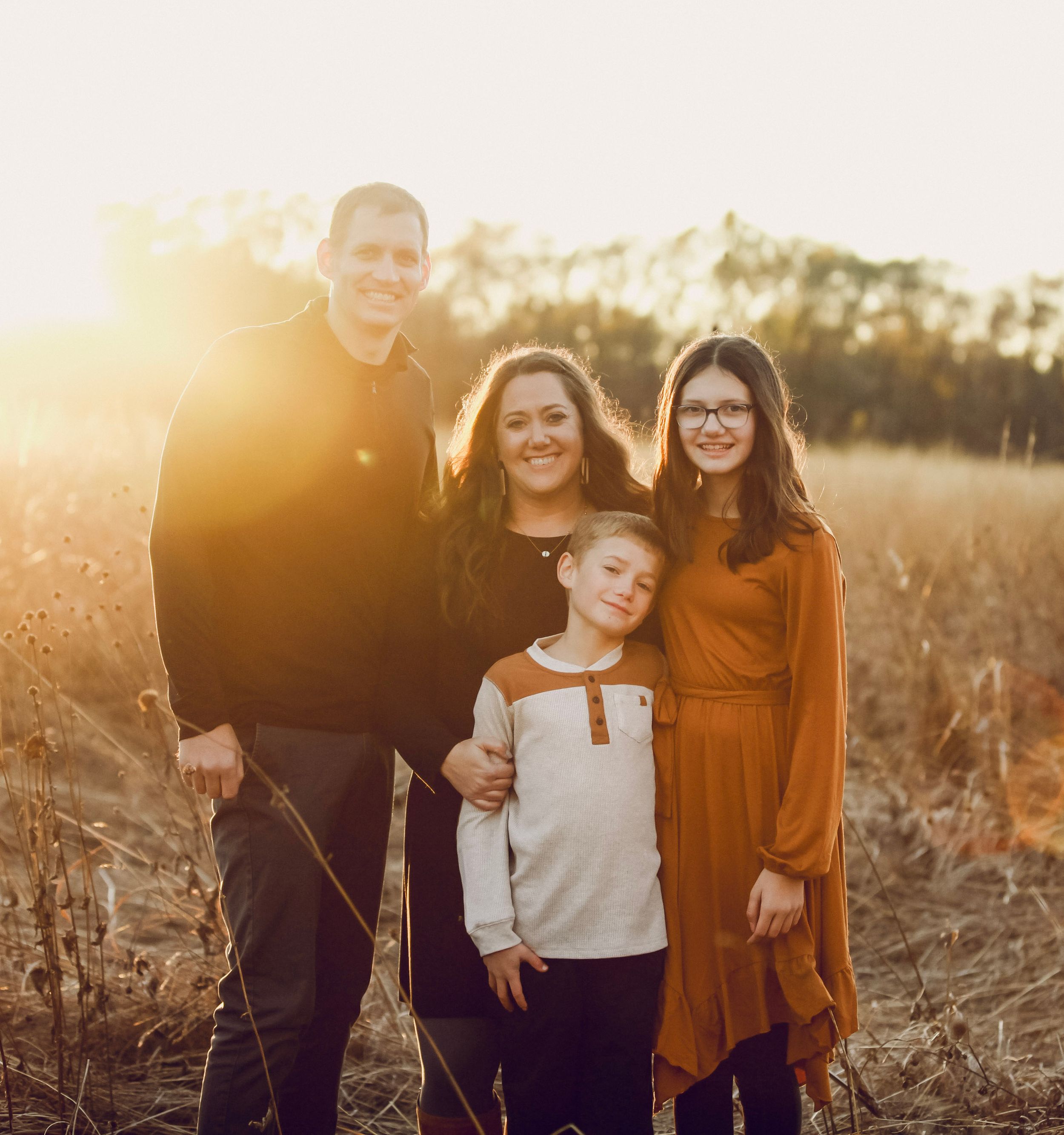 Family of four smiling together in a golden sunlit field during autumn, wearing casual fall outfits.