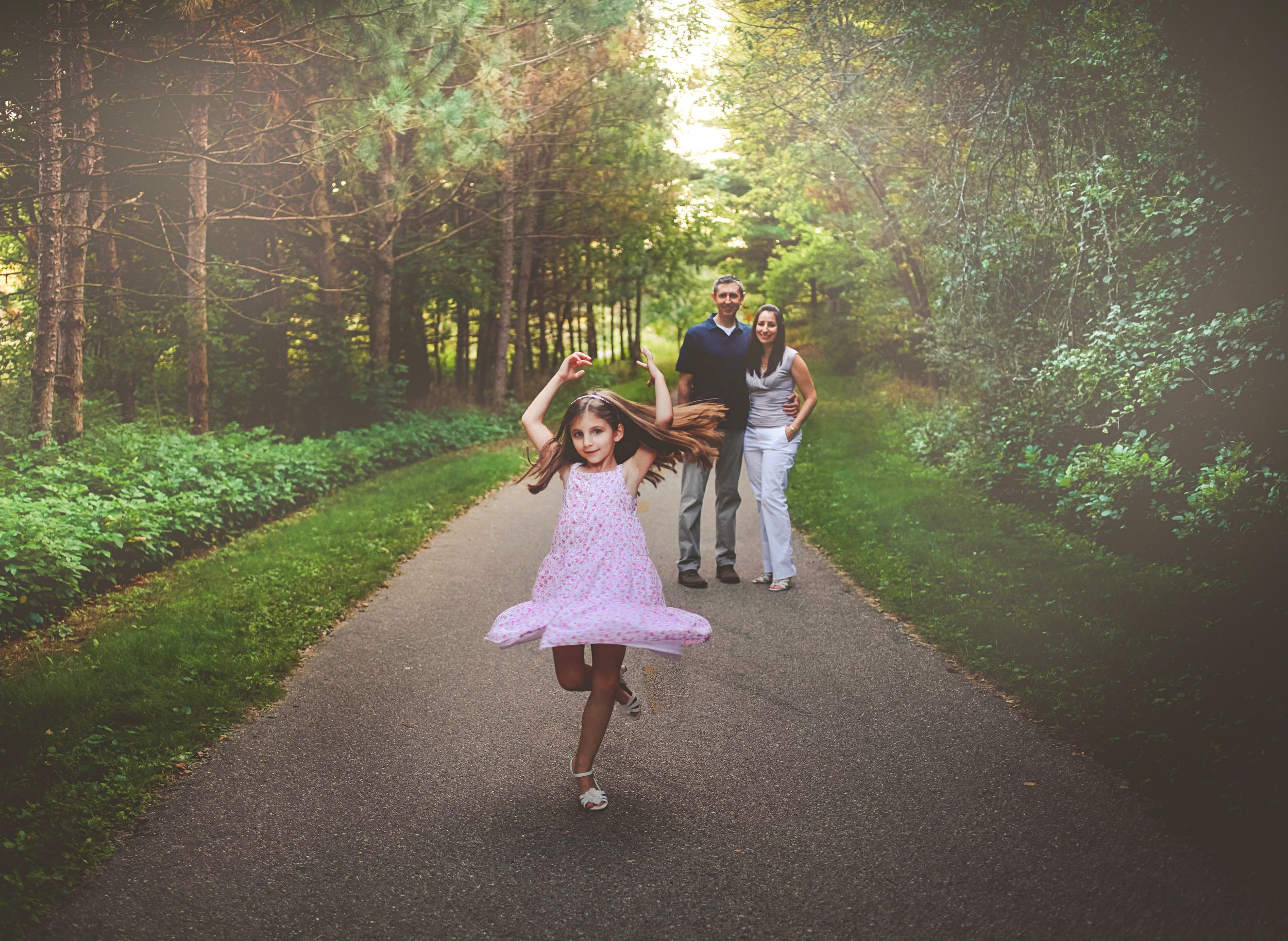 A joyful girl in a pink dress twirls on a forest path while her parents smile in the background.