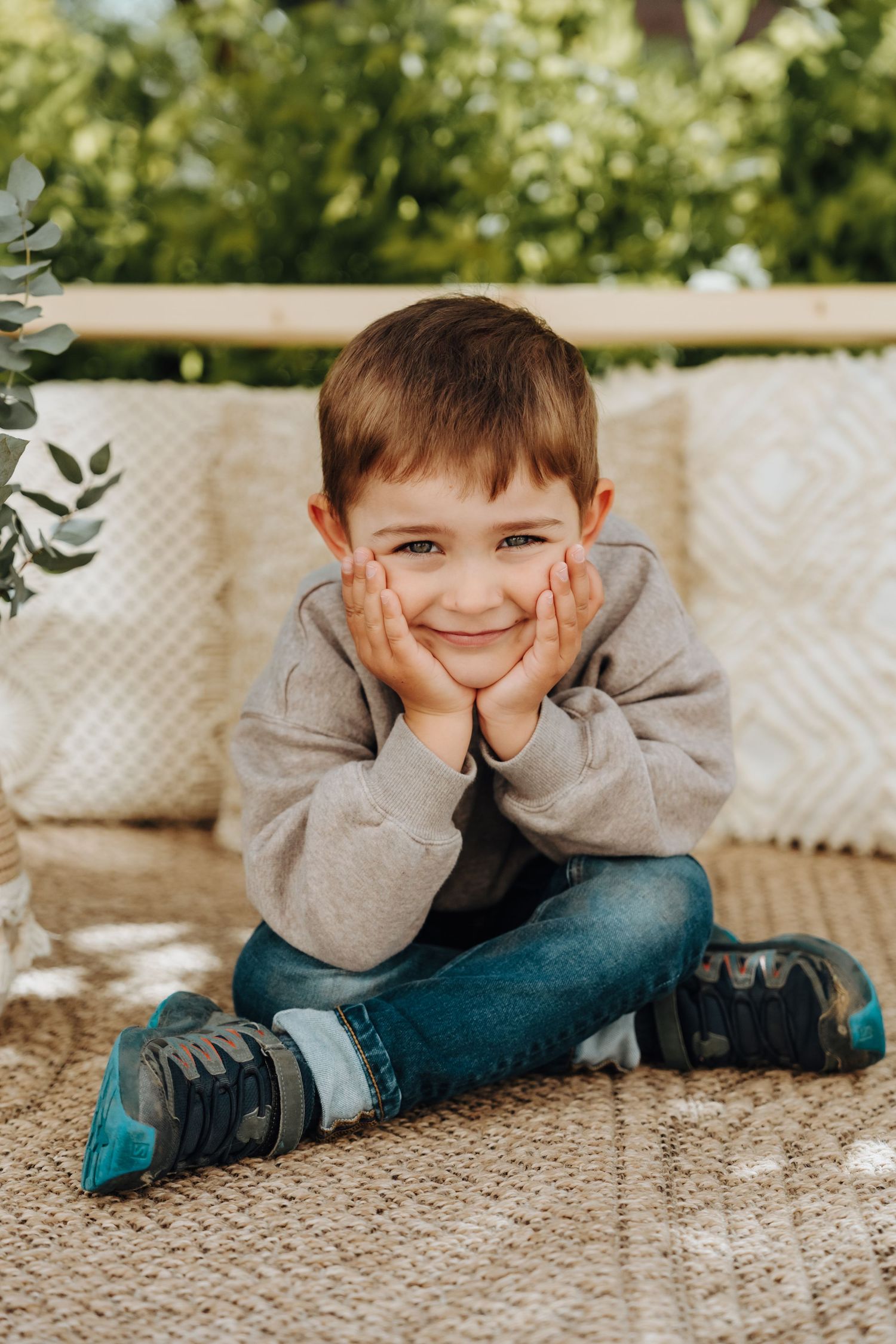 Smiling young boy sitting cross-legged outdoors, resting chin on hands, wearing gray hoodie and jeans.