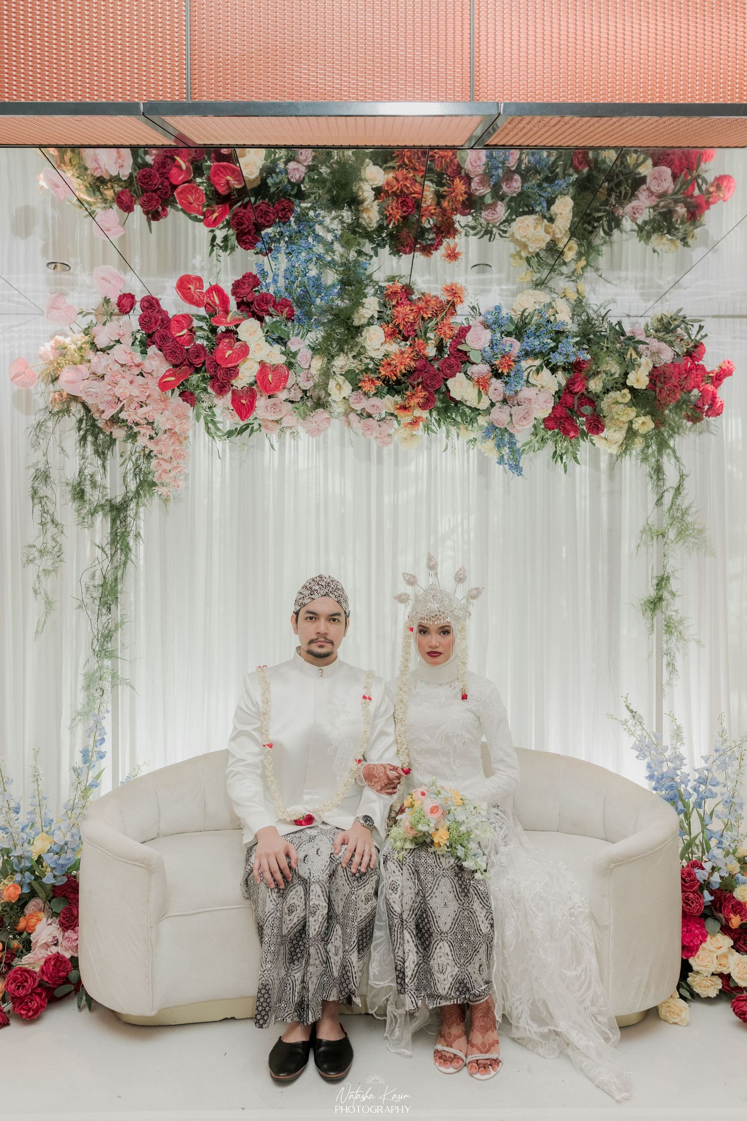 Muslim couple walking hand in hand in a lush garden, woman in hijab and white dress, man in cream jacket.