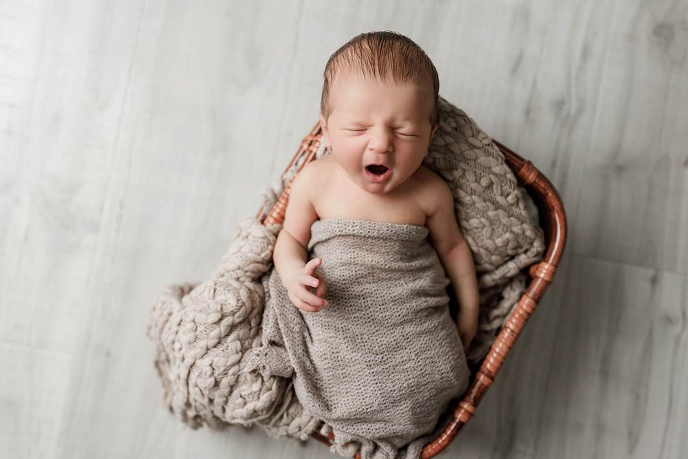 Newborn baby yawning while wrapped in a cozy gray blanket inside a wicker basket.
