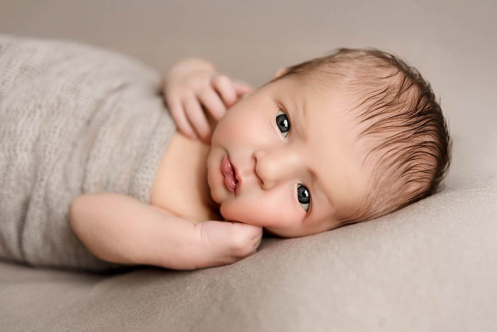 Serene newborn baby with dark hair lying on a soft gray blanket in a professional studio portrait.