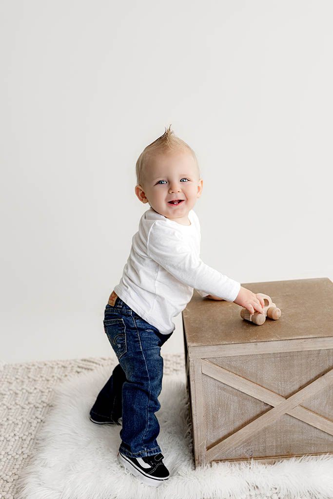 Smiling toddler boy in white shirt and jeans stands beside a wooden crate, holding a toy car.