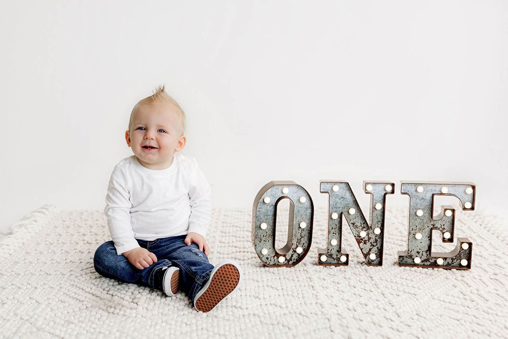 Happy baby boy celebrating his first birthday sitting next to a decorative ONE sign on a white backdrop.