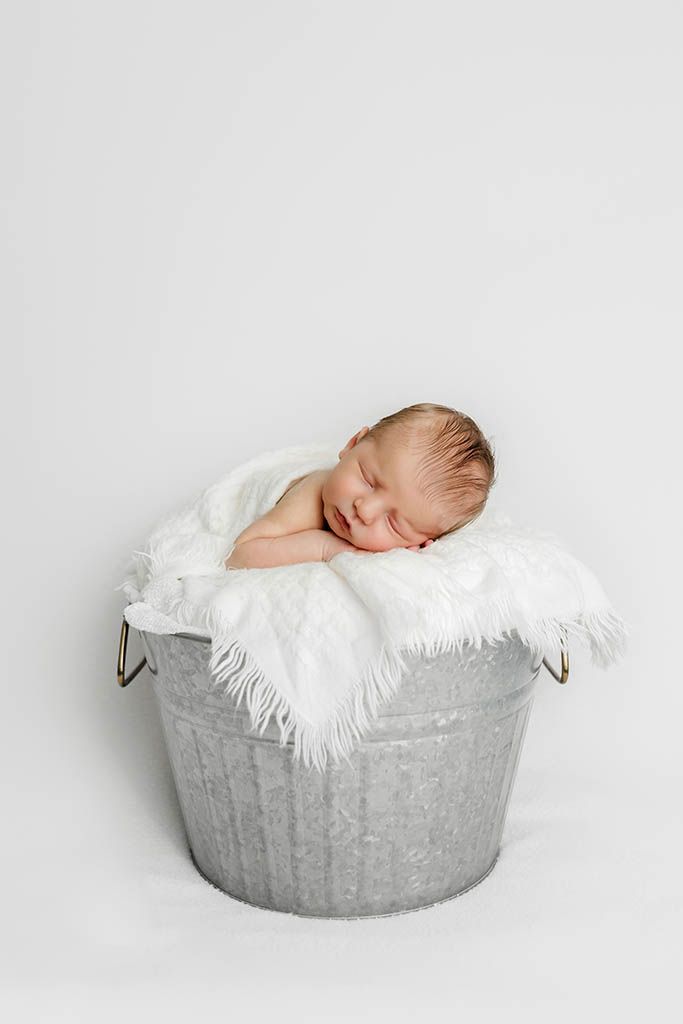 Sleeping newborn baby resting peacefully in a rustic metal bucket lined with soft white fur, on white background.