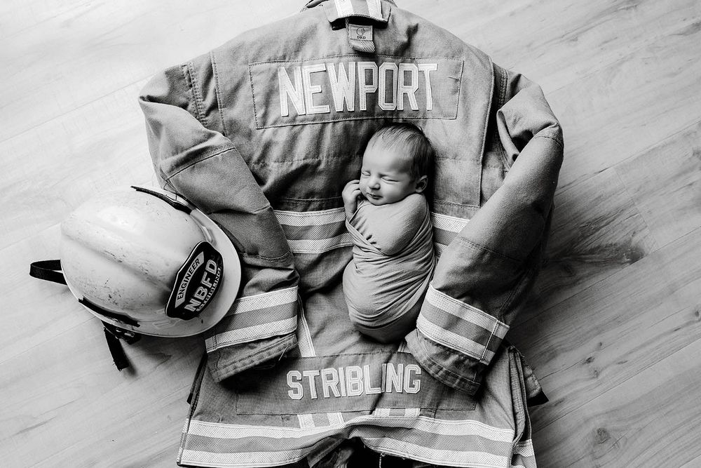 Newborn baby swaddled on Newport Stribling firefighter turnout gear with helmet in black and white photo.