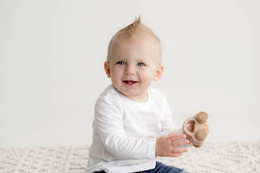 Smiling baby boy in white long sleeve shirt holding a wooden toy while sitting on a white textured blanket.
