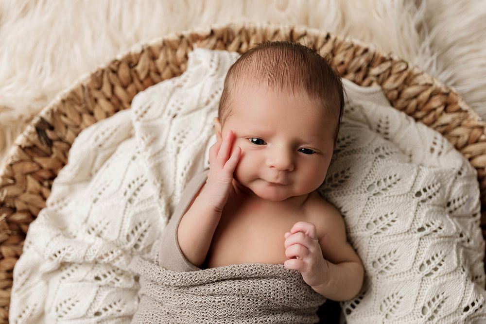 Adorable newborn baby wrapped in knit blanket posed in woven basket with soft white fur background.