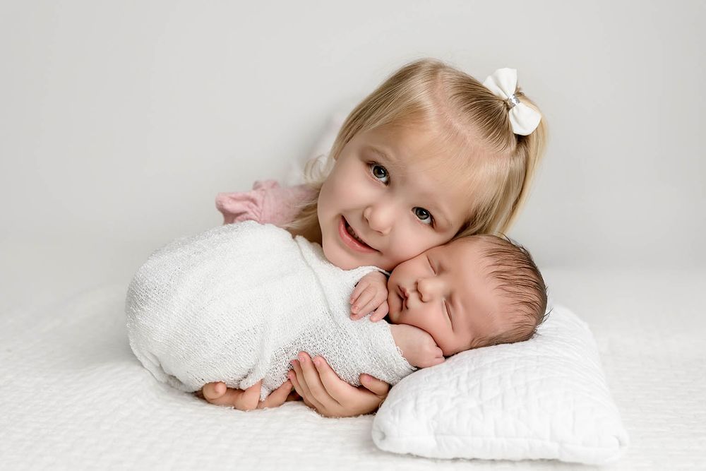 Smiling toddler girl with white bow hugs sleeping swaddled newborn baby on white backdrop.